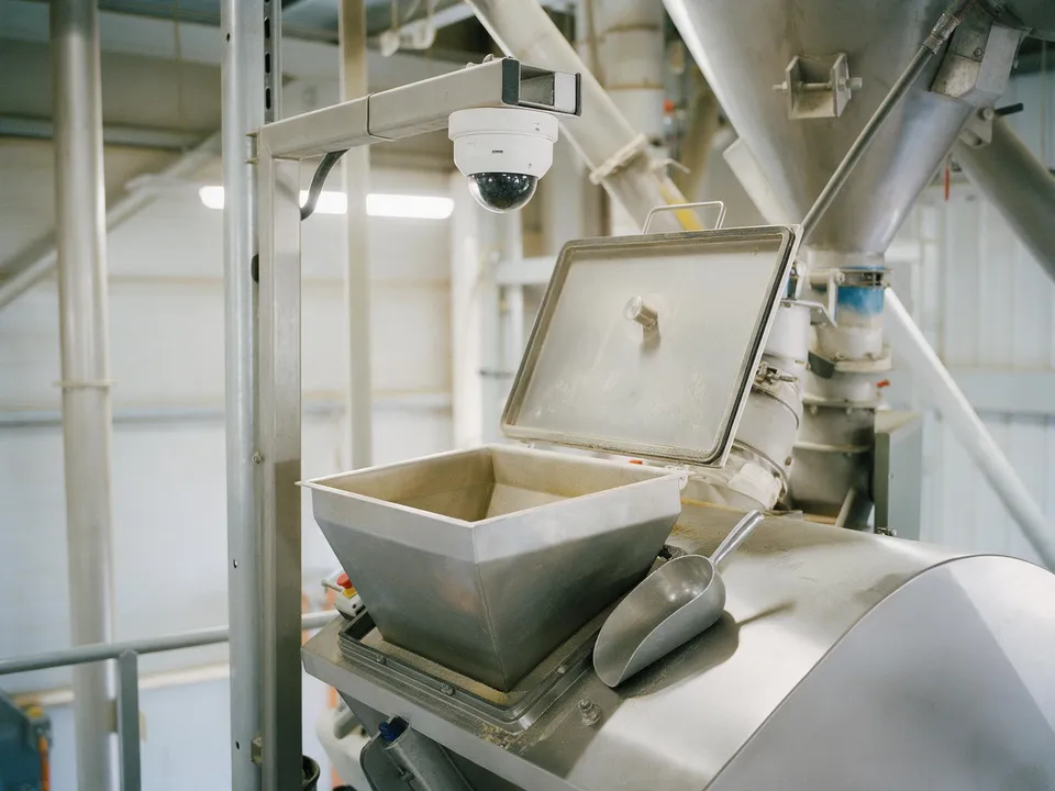 An open stainless steel hand-addition hopper at a mixer inlet with a scoop beside it and a dome camera above